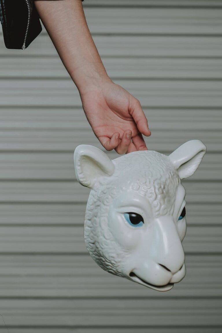 A hand holding a white sheep mask by its ear against a textured background.